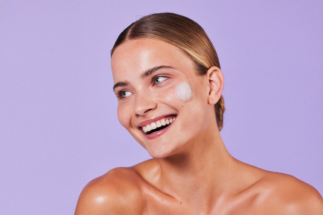 Smiling teen with clear skin and a foamy cleanser on her cheek, set against a lavender background – representing gentle acne care.